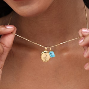May include: A gold necklace with a delicate chain. The necklace features two pendants: a round locket with a floral design and a rectangular, light blue gemstone. The locket is engraved with a flower.