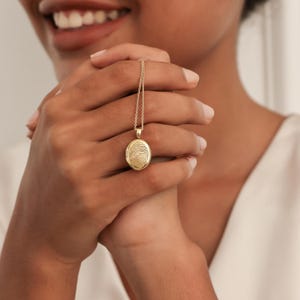 May include: A gold-colored oval pendant necklace with a fingerprint design. The pendant hangs from a delicate chain. The necklace is held in a person's hands, with the focus on the jewelry.