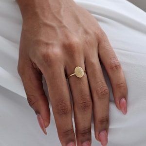 May include: A gold ring featuring a textured oval design. The ring is shown on a hand with manicured nails, set against a white background. The ring's band is thin, and the oval is the focal point.