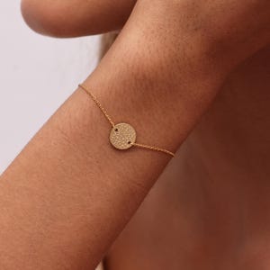 May include: A gold-toned bracelet featuring a delicate chain and a central circular charm. The charm has a textured, hammered surface finish. The bracelet is secured around the wrist with a thin, polished metal link chain design.