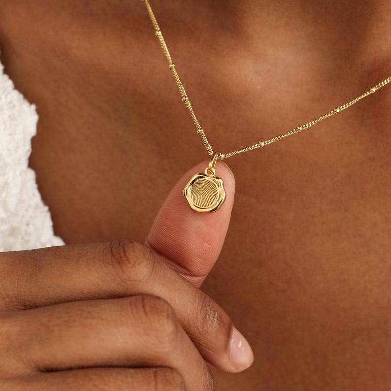 May include: A gold-colored necklace with a fingerprint pendant. The pendant is round with a detailed fingerprint design. The necklace is on a person's neck and finger.
