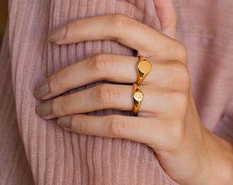 May include: Two gold-toned rings worn on adjacent fingers. The upper ring features a circular, textured face, while the lower ring displays a small, engraved paw print design on its flat, circular surface. Both rings have a polished finish.