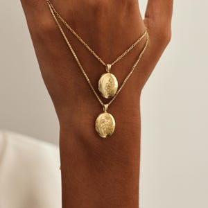 May include: Two gold-colored necklaces with oval lockets. Each locket features a floral design. The necklaces are displayed on a hand, showcasing the jewelry's delicate chains and intricate details. The lockets are likely for holding small photos or keepsakes.