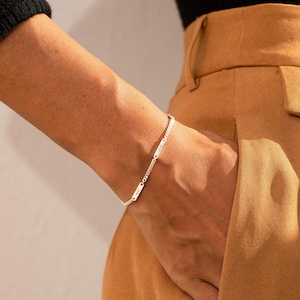 May include: A silver-toned chain bracelet featuring two small, rectangular metal plates. Each plate is engraved with text, and the bracelet is constructed with a delicate curb chain link design that wraps around the wrist area.