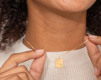 May include: A gold-toned necklace featuring a thin chain and a small, rectangular pendant. The pendant has a smooth, polished surface with a tiny, engraved caduceus medical symbol positioned in the lower right corner of the rectangular charm.