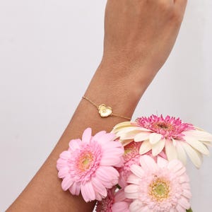 May include: A delicate gold-toned chain bracelet featuring a small, central heart-shaped charm with an engraved floral design. The bracelet is worn on a wrist, positioned near a cluster of vibrant pink and white gerbera daisy flowers.