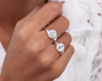 May include: Two polished silver signet rings worn on adjacent fingers. Each ring features a flat, circular face engraved with a delicate, black-lined botanical illustration of a single flower stem with leaves and small buds.