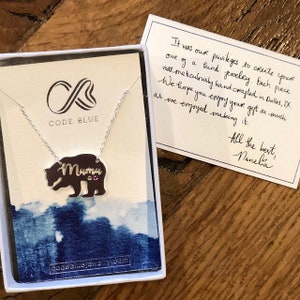 May include: A silver necklace with a bear pendant that says "Muma" with two pink gemstones. The necklace is in a white box with a blue and white design. The box also includes a handwritten note that says "It was our privileges to create your one of a kind jewelry. Each piece was meticulously hand crafted in Dallas, TX. We hope you enjoy your gift as much as we enjoyed making it. All the best, Nanela."