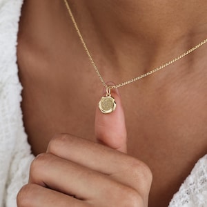 Könnte beinhalten: Eine goldfarbene Halskette mit einem kleinen, runden Anhänger. Der Anhänger hat eine strukturierte Oberfläche und hängt an einer filigranen Kette. Die Halskette wird vor einem weißen Hintergrund getragen.