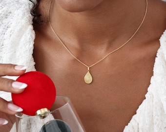 Puede incluir: Un collar con colgante dorado con forma de lágrima con diseño de huella dactilar. El collar lo lleva una persona con un top de encaje blanco. Una bola roja de adorno se sostiene en la mano.