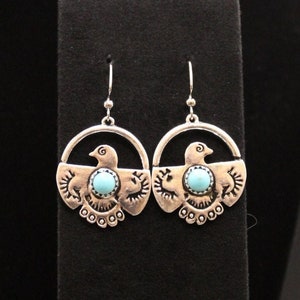 May include: Silver dangle earrings with a turquoise stone in the center of a bird design. The bird is surrounded by a circle with decorative details.