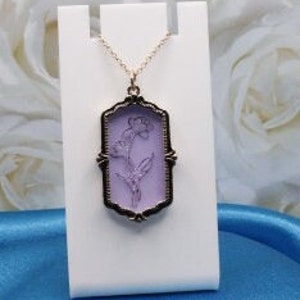 May include: A delicate necklace featuring a lavender-colored floral pendant. The pendant is encased in a black rectangular frame with ornate detailing and hangs from a gold-colored chain. The necklace is displayed on a white stand against a soft blue backdrop.
