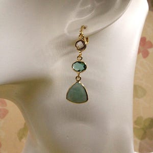 May include: Gold dangle earrings with three green and pink gemstone drops. The bottom drop is a triangular shaped stone.
