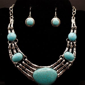May include: A silver-toned metal necklace and earring set featuring five turquoise-colored stones with dark veining. The necklace has a wide, multi-strand design with a large central stone and matching round drop earrings with hook backings.