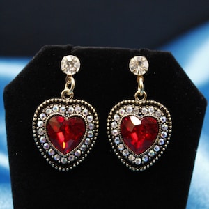 May include: A pair of heart-shaped earrings with red crystal centers surrounded by a halo of smaller clear crystals. The earrings are on a black display stand.