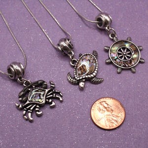 May include: Three silver-toned necklaces with ocean-themed pendants: a crab, a turtle, and a ship's wheel. Each pendant has iridescent abalone shell accents. A penny is included for size reference. The necklaces are on a purple glittery surface.