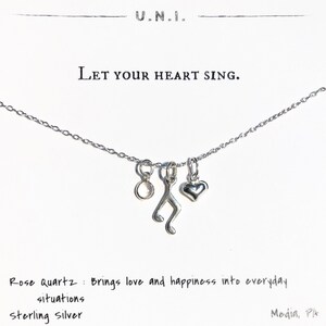 May include: A silver necklace with a music note charm, a heart charm, and a rose quartz charm. The necklace is on a white background with the text "Let your heart sing." and "Rose Quartz: Brings love and happiness into everyday situations sterling silver".