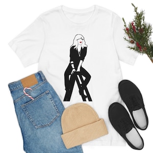 Op de afbeelding: Wit T-shirt met een zwart-witte illustratie van een vrouw met blond haar die op een ladder zit. De vrouw draagt een zwart shirt en een zwarte broek.
