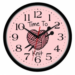 May include: A round wall clock with a black frame and a light pink face. The clock features black numbers from 1 to 12, black hour and minute hands, and a red second hand. In the center is a pink yarn ball shaped like a heart with two knitting needles.