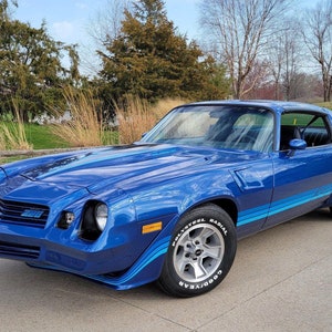 Op de afbeelding: Een blauwe Chevrolet Camaro uit de jaren 70 met een zwarte streep en blauwe race strepen. De auto heeft een zwarte grille en een zwarte bumper. De auto staat geparkeerd op een geplaveide oprit.