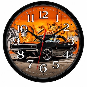 May include: A round wall clock with a black frame featuring a black vintage muscle car parked in front of an orange graffiti-covered wall. The clock face has large white serif numerals from one to twelve, black hour and minute hands, and a red second hand.