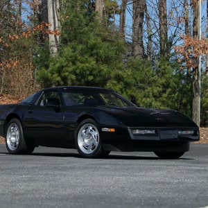 May include: A black sports car with silver wheels is parked on a paved surface. The car has a sleek, aerodynamic design, with a low profile and a sloping hood. The background features trees and a clear sky.