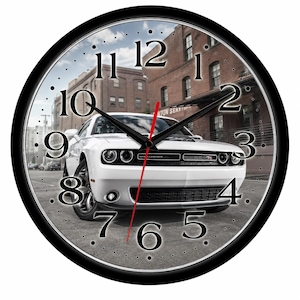 May include: A round wall clock with a black frame featuring a white Dodge Challenger car parked on a city street in front of brick buildings. The clock face has large black serif numbers from 1 to 12, black hour and minute hands, and a thin red second hand.