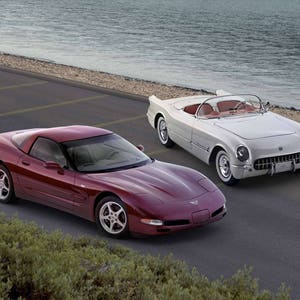 May include: Two classic sports cars are parked on a paved road near a body of water. One is a maroon coupe, and the other is a white convertible with a red interior. The cars are positioned side-by-side.