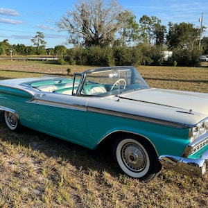 May include: A turquoise and white 1957 Ford Fairlane 500 Skyliner convertible. The car is parked on a grassy field with a blue sky in the background.