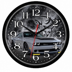May include: A round wall clock with a black frame featuring a gray Dodge Challenger SRT Demon car graphic on a smoky background. The clock face displays white Arabic numerals, black hour and minute hands, and a thin red second hand.
