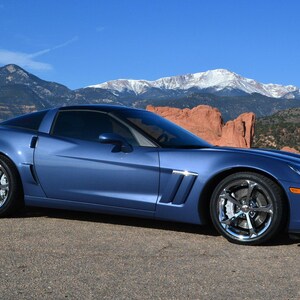 May include: A blue sports car with chrome wheels is parked on a paved road. The car has a sleek design and is set against a backdrop of mountains and a clear blue sky. The car's paint has a metallic sheen.
