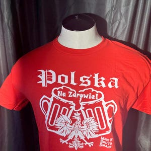May include: Red t-shirt with a white graphic of two beer mugs clinking, a Polish eagle, and the text "Polska Na Zdrowie!" and "Made in South Omaha".