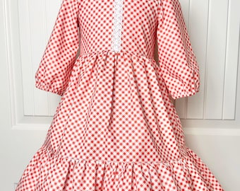 May include: A red and white gingham dress with three-quarter sleeves and a ruffled hem. The dress features a white lace detail down the front. The dress is displayed on a mannequin. The dress is suitable for a child.