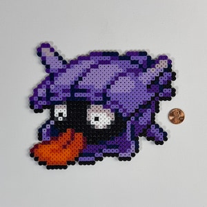 May include: A pixel art design of a cartoon character, primarily purple with black and orange accents. The character has a large, stylized head with a prominent beak and eyes. A penny is next to the character for size comparison.