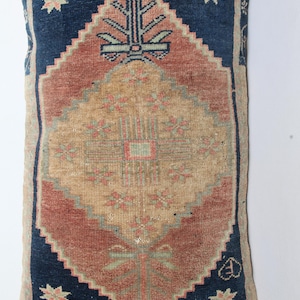 Puede incluir: Cojín rectangular con diseño de alfombra vintage. El cojín presenta un patrón central de diamante en beige y rojo, enmarcado por un borde azul marino con acentos en forma de estrella. El diseño evoca una estética tradicional y artesanal.