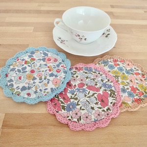 Peut inclure: Trois dessous-de-verre en crochet avec des motifs floraux. Les dessous-de-verre sont en bleu, rose et marron.