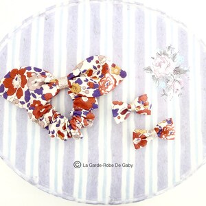 May include: A set of three hair scrunchies with a floral pattern in shades of orange, purple, and white. The scrunchies are made of fabric and have a gathered look. The scrunchies are on a striped background.