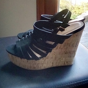 Puede incluir: Sandalias de cuña negras con suela de corcho. Las sandalias tienen múltiples correas en el pie y un cierre de hebilla alrededor del tobillo. La suela de plataforma es de corcho marrón claro.