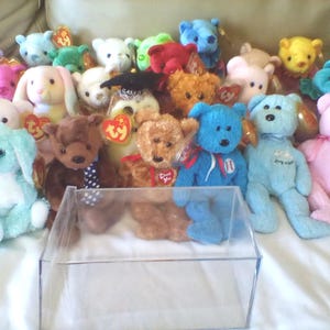 May include: A collection of colorful plush toys, including bears and bunnies, in various colors such as pink, blue, and brown. Some toys have tags attached. A clear plastic display case is in the foreground.