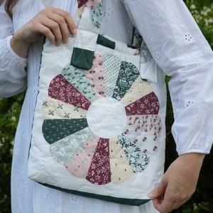 May include: A white quilted tote bag with a green fabric handle and a patchwork design featuring a circle of colorful fabric pieces. The bag is made of cotton and has a quilted texture.