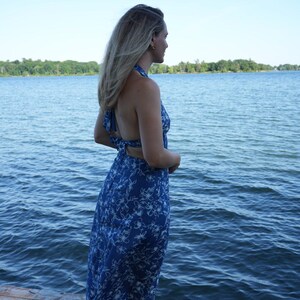 May include: A blue halter maxi dress with a white floral pattern. The dress has an open back. The person is standing on a wooden dock. The background features a lake and a clear blue sky.