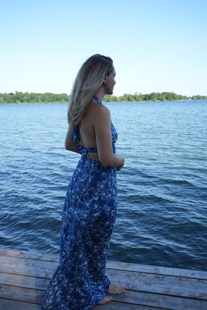 May include: A blue halter maxi dress with a white floral pattern. The dress has an open back. The person is standing on a wooden dock. The background features a lake and a clear blue sky.