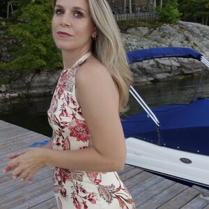 May include: A woman in a halter-neck dress with a floral design in red, pink, and brown hues. The dress is set against a backdrop of a wooden dock, water, and trees. A boat with a blue cover is visible.