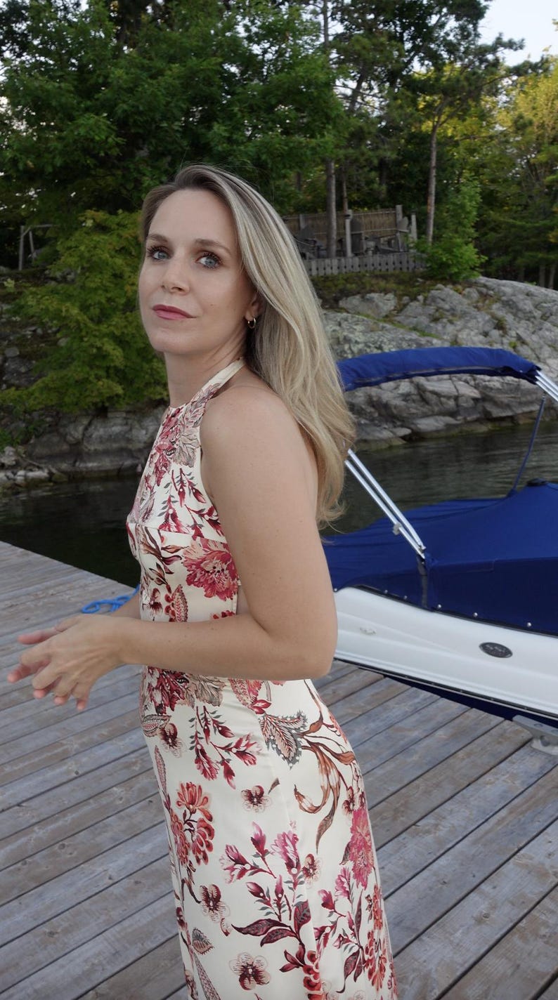 May include: A woman in a halter-neck dress with a floral design in red, pink, and brown hues. The dress is set against a backdrop of a wooden dock, water, and trees. A boat with a blue cover is visible.
