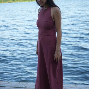 May include: A burgundy halter-neck maxi dress. The dress features a fitted bodice and a flowing, wide-leg design. The model is standing on a wooden dock with a lake in the background. The dress is made of a soft, flowing fabric.