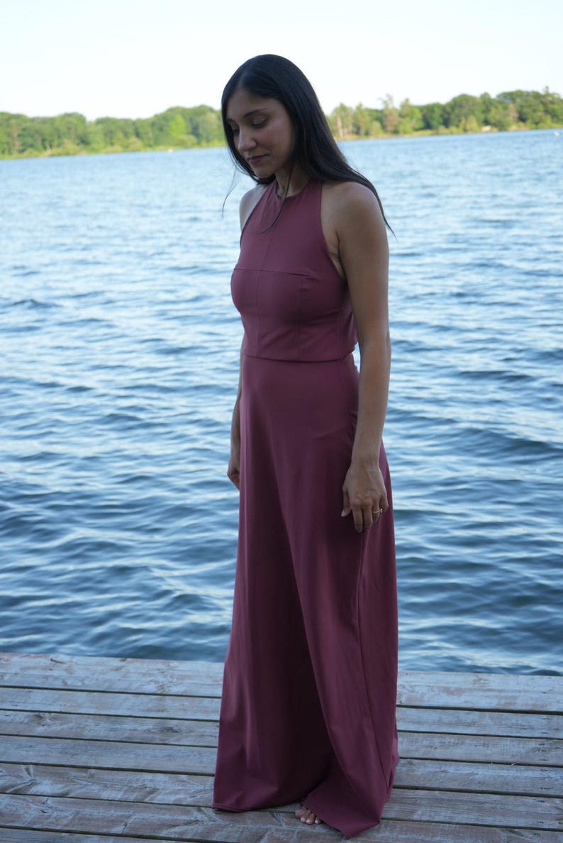 May include: A burgundy halter-neck maxi dress. The dress features a fitted bodice and a flowing, wide-leg design. The model is standing on a wooden dock with a lake in the background. The dress is made of a soft, flowing fabric.