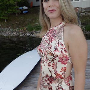 May include: A cream-coloured halter dress with a floral pattern in shades of red and pink. The dress has a fitted bodice and a flowing skirt. The model is standing on a wooden dock near a lake.