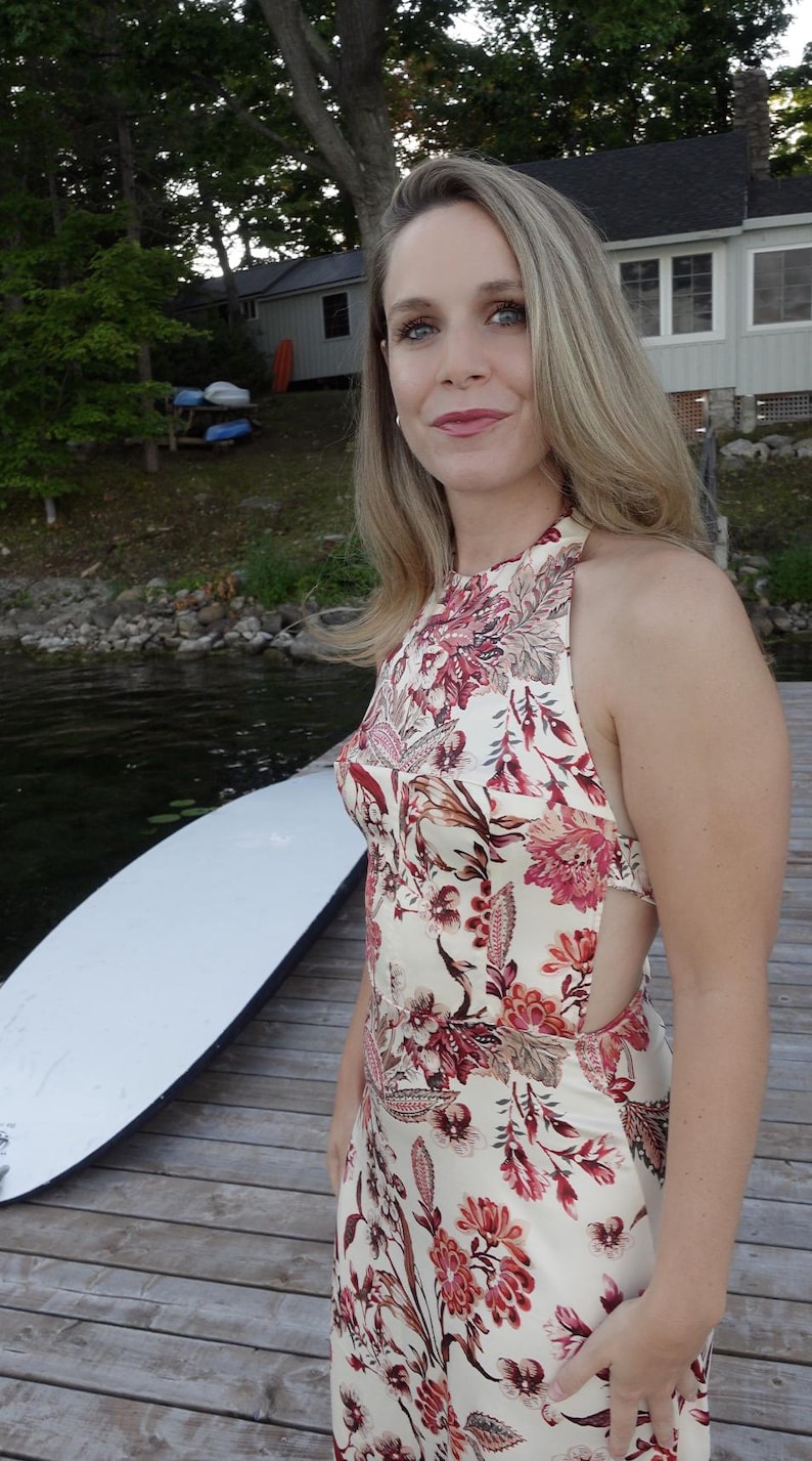 May include: A cream-coloured halter dress with a floral pattern in shades of red and pink. The dress has a fitted bodice and a flowing skirt. The model is standing on a wooden dock near a lake.