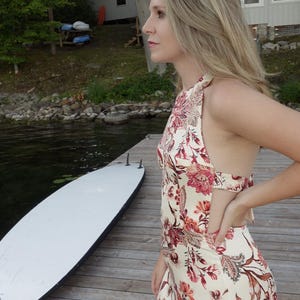 May include: A cream halter dress with a floral pattern in red and brown. The dress is worn by a person standing on a wooden dock next to a white surfboard. The background includes a lake and trees.