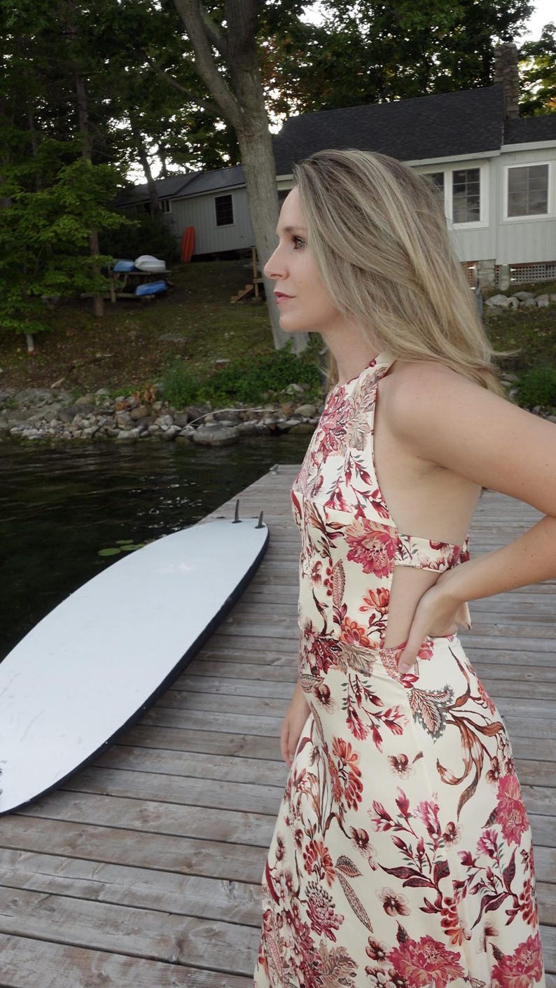 May include: A cream halter dress with a floral pattern in red and brown. The dress is worn by a person standing on a wooden dock next to a white surfboard. The background includes a lake and trees.